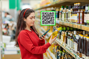 A pretty Caucasian woman scans the qr code on a bottle of oil using her smartphone. Qr code icon above the product. In the background is a supermarket. Concept of modern technologies and shopping