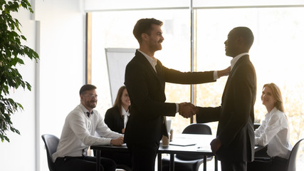 Young male team leader shaking hands with african american employee, congratulating with good job...