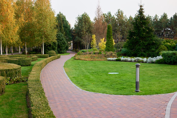 Empty city park landscape. Paving stone walkway, green lawn, bushes, and autumnal trees.