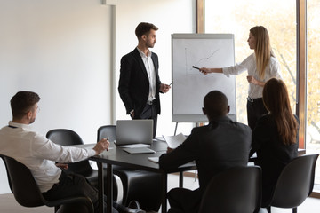 Multiracial team of employees sitting at table, listening to male and female presenters, explaining financial growth company strategy. Young woman speaker leader showing sales plan to diverse workers.