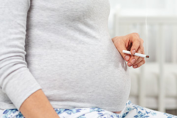 Pregnant woman with a cigarette in her hand, sitting on the bed. Hands with a cigarette and belly close-up. Baby cradle in the background. The concept of nicotine addiction and women's health