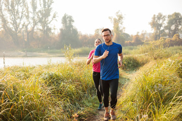 Young caucasian man and woman running along the trail trough tall grass by the river in early morning