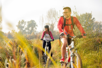 Happy young caucasian man and woman riding mountain bikes along the trail through tall grass in early morning