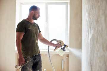 Side view of man painting with airbrush, apartment repairment against small window in the daytime. Man with a beard is dressed in paint-smeared T-shirt. Repair concept