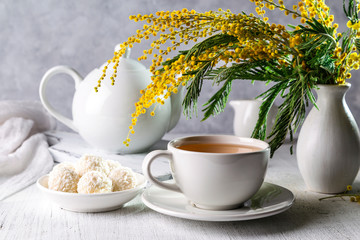 A bouquet of mimosas in a vase with tea and candies on a light background.