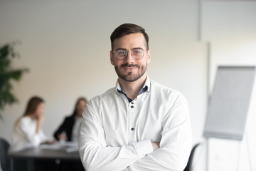 Confident happy young male manager in eyeglasses standing with folded hands head shot portrait. Smiling company owner, startup businessman, team leader, successful employee looking at camera. - obrazy, fototapety, plakaty