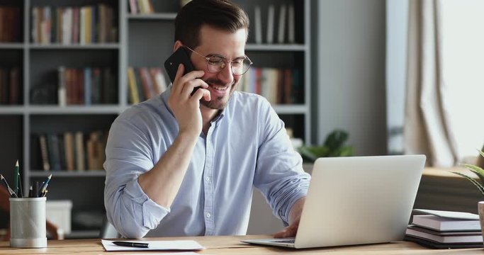 Smiling business man using laptop talking on cell phone sits at desk. Happy confident male professional manager web designer consulting client about online project making business call at workplace.