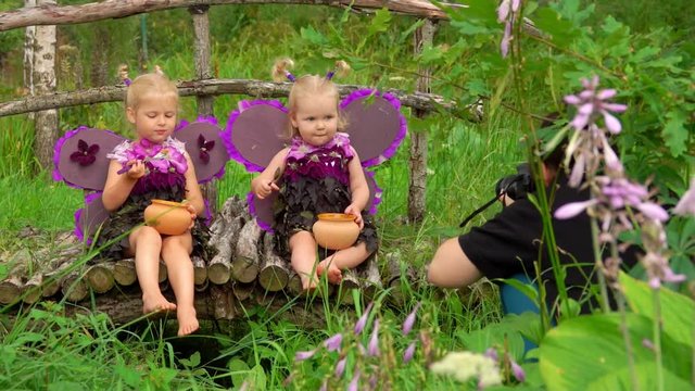 Female photographer takes pictures of beautiful girls playing violet butterflies. Girls are eating honey and wearing handmade butterfly wings