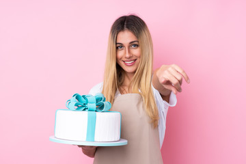 Young Uruguayan pastry woman with a big cake over isolated pink background points finger at you with a confident expression