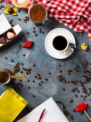 life style composition with cup of coffee on grey cement background, red tablecloth, trendy sweets, party lips, yellow mockup pack, notebook, brown beans, copy space in center. Vertical, Top view