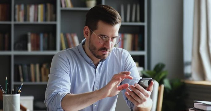 Male millennial professional holding modern smartphone texting message in office. Young businessman using helpful mobile apps for business time management organization concept sitting at work desk.