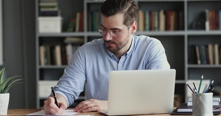 Focused business man working on laptop computer making notes sitting at home office desk. Young adult male professional manager doing marketing research writing checklist on paper at workplace. - Powered by Adobe