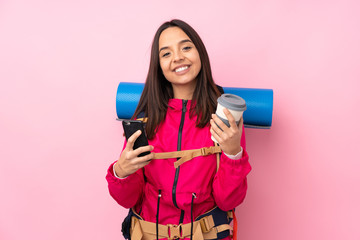Young mountaineer girl with a big backpack over isolated pink background holding coffee to take away and a mobile