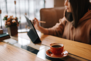 Close-up of a cup of dark coffee on a table in a cafe. In the background sits a young woman in casual clothes and works at a digital tablet.