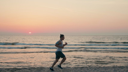 Sporty Hadsome Man is Jogging on Beach with Headphones, Running along Ocean Coast at Sunset or Sunrise