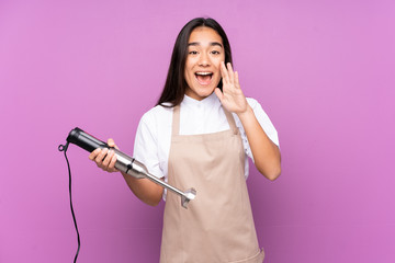 Indian woman using hand blender isolated on purple background shouting with mouth wide open
