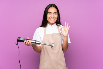 Indian woman using hand blender isolated on purple background showing an ok sign with fingers