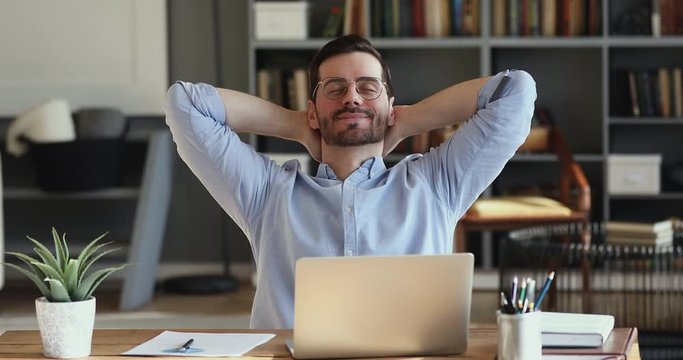 Relaxed businessman takes break to relieve stress. Satisfied happy male employee meditating sitting at workplace desk holding hands behind head. Office worker finished work feels peace of mind concept