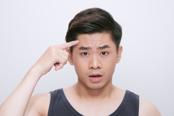 Young asian chinese man wearing t-shirt standing over isolated white background pointing unhappy to pimple on forehead, ugly infection of blackhead. Acne and skin problem