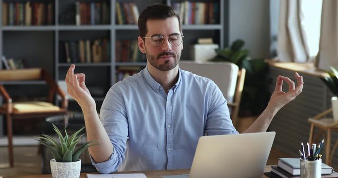 Mindful young businessman meditating in office room managing work stress. Calm serene man taking break seated at home office desk doing yoga breathing exercise. Peace of mind at workplace concept.