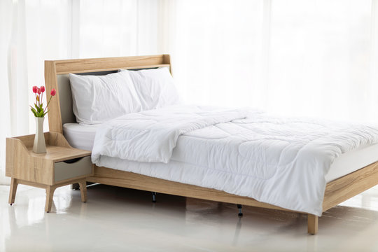 The wooden bed is laid with white sheets in empty room surrounded by white curtains and soft sun light .