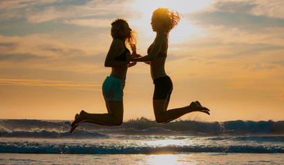 Silhouette of two beautiful women running and jumping on the beach shore with waves in the background at sunset or sunrise.