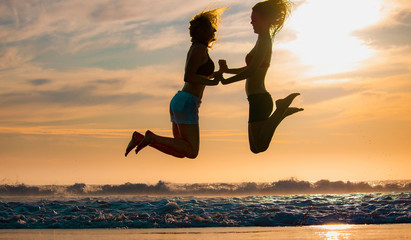 Silhouette of two beautiful women running and jumping on the beach shore with waves in the background at sunset or sunrise.