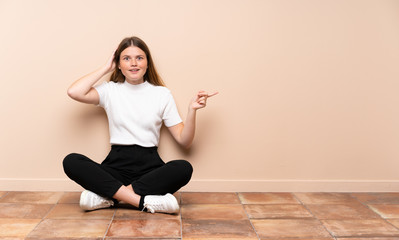Fototapeta na wymiar Ukrainian teenager girl sitting on the floor surprised and pointing finger to the side