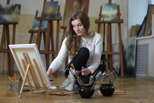 Woman artist sits on the floor among easels with paintings in an art studio and paints an oil painting on canvas.