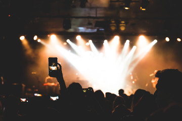 A person is taking a picture with her phone in a concert. The people enjoying with music in the party. Selective focus.
