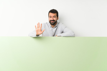 Young handsome man with beard holding a big green empty placard counting five with fingers