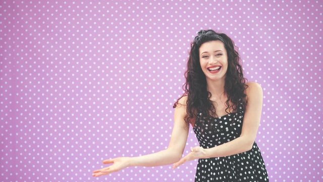 Lovely young girl with kinky hair, in black polka-dots dress dancing, waving comically her hands, pointing at copy space for text or product.