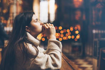 A young woman is sitting with her hands folded and is praying in a church - obrazy, fototapety, plakaty