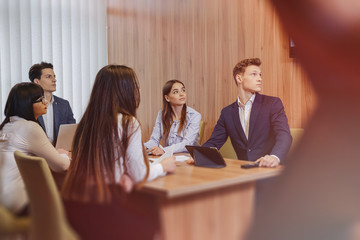 large team of people is working at one table for laptops, tablets and papers, on the background a large TV set on a wooden wall