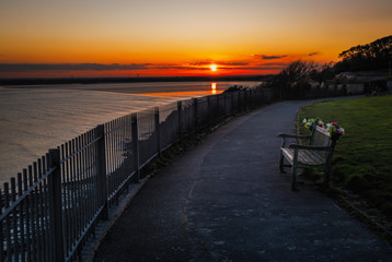 Sunset reflecting in the sea with the cliff top promenade and memorial bench in the foreground in...
