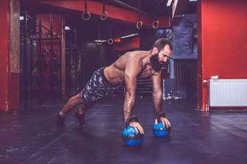 Young muscular bearded man with naked torso doing push ups with kettlebell weights