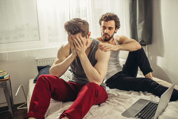 offended young caucasian gay sit with closed face, his partner soothing and support him. homosexual married couple after first wedding night. disagreement in homosexual family - Powered by Adobe