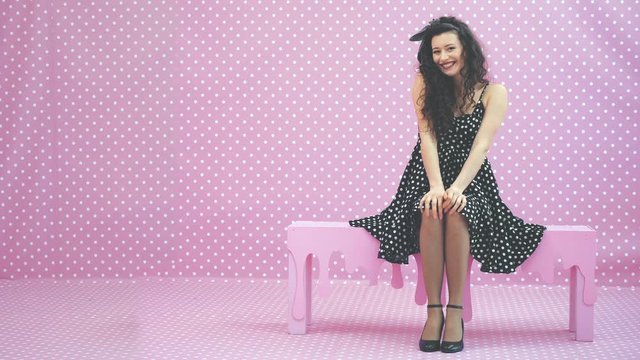 Kinky brunnete girl in beautiful dress sitting on the pink bench, blowing an airkiss. Polka-dots.
