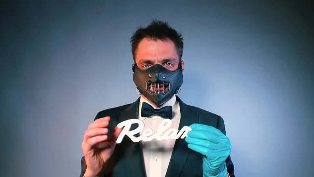 A man in a suit and mask at a party holds the word relax in his hand. The second hand is wearing a glove. The concept of a good time.