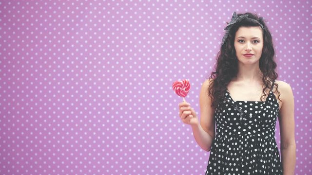 Lovely young curly girl standing, holding sweet red lollipop on the stick in her hand, petting her belly, looking at the candy, showing tisk.