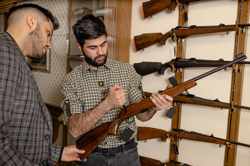young caucasian customer and salesman discussing characteristics of rifle in guns store. weapon,...