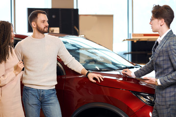 young couple thinking about buying a car, close up photo. dealer and clients standing in front of the red car at dealership. close up photo