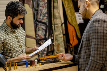 confident caucasian salesman checking document of customer, license to use weapon in guns store, young man came to get rifle for hunting