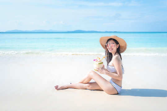 beautiful young tan skin asian woman sitting on the beach wearing bikini ,beach hat, sunglasses smiling with coconut drink. relaxing summer holiday with sunny blue sky, vacation concept