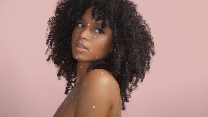 Mixed race black woman with curly hair covered by crystal makeup on pink background in studio...