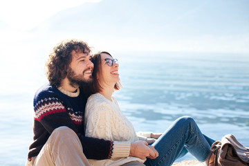 Couple in love hugging and sitting at winter sea beach outdoors. Young travelers smiling and...