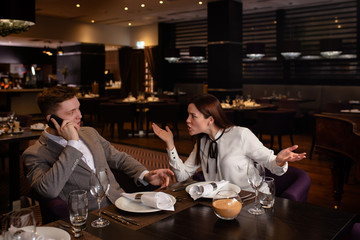 bad evening in a restaurant. young caucasian couple quarreled in representative luxury restaurant, they don't talk with each other. woman scream at man talking on phone