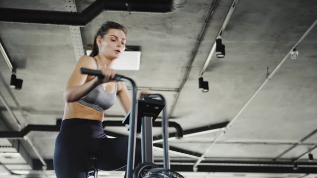 Tracking from below medium shot of determined young woman practicing exercise on air resistance stationary bike in gym