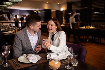 young caucasian couple have dinner together, look at smartphone in restaurant. happy woman show cool video to boyfriend. indoors. man and woman enjoy spending time together