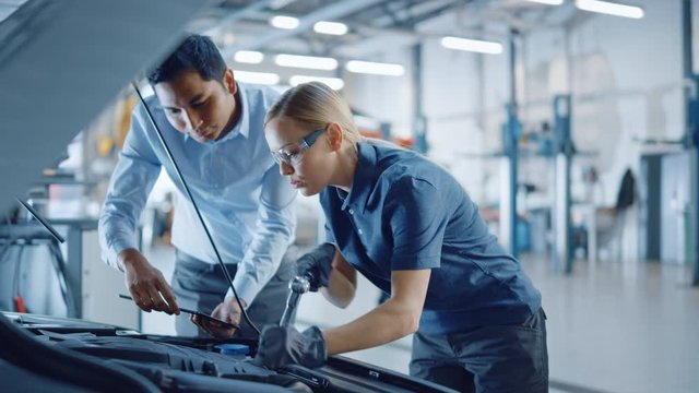 Instructor with a Tablet Computer is Giving a Task for a Future Mechanic. Female Student Inspects Car Engine and Uses Ratchet. Assistant is Checking Cause of Breakdown in Vehicle in a Car Service. 